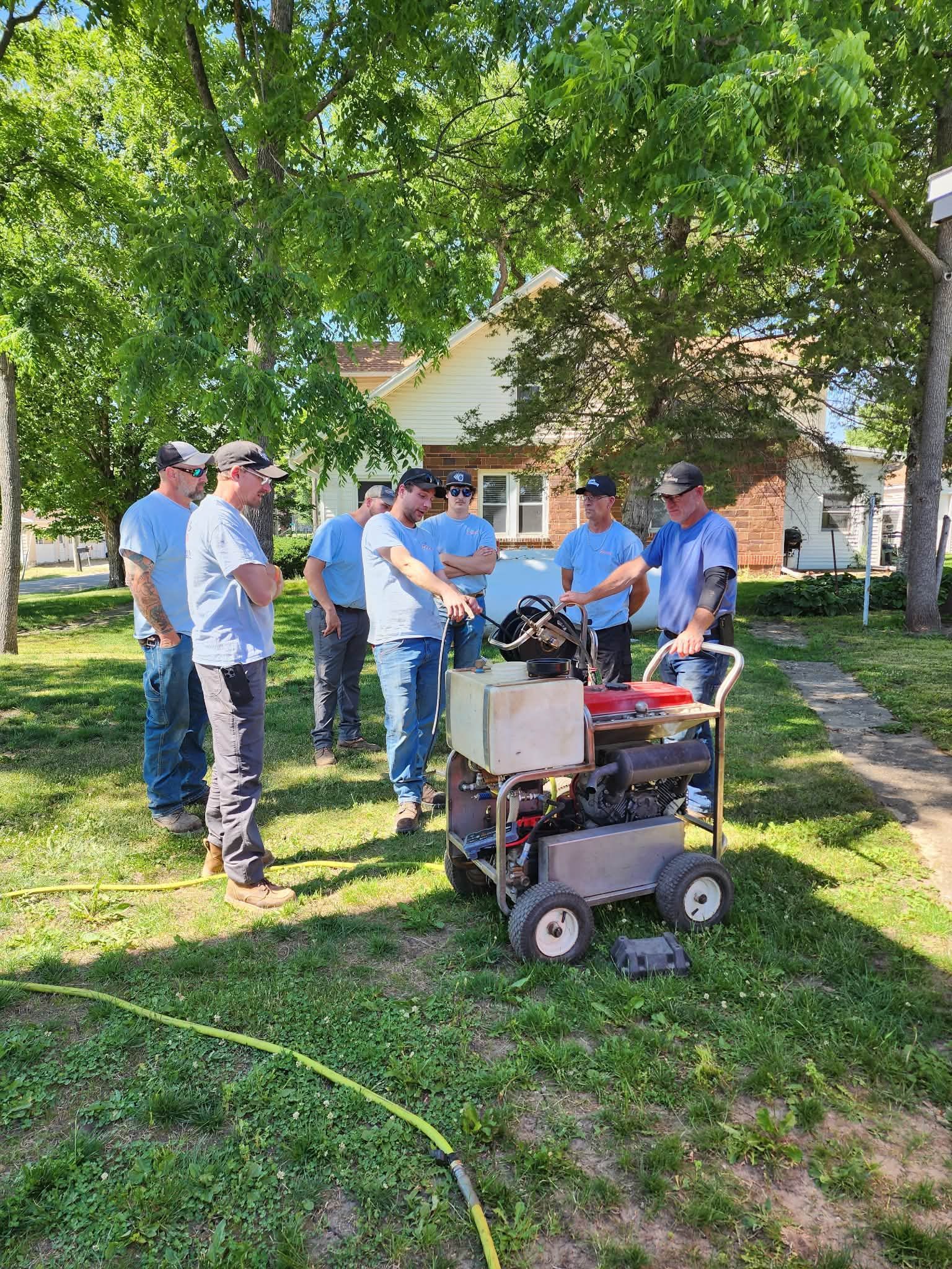 People gathered around a cart and equipment under a large tree in a sunny yard.