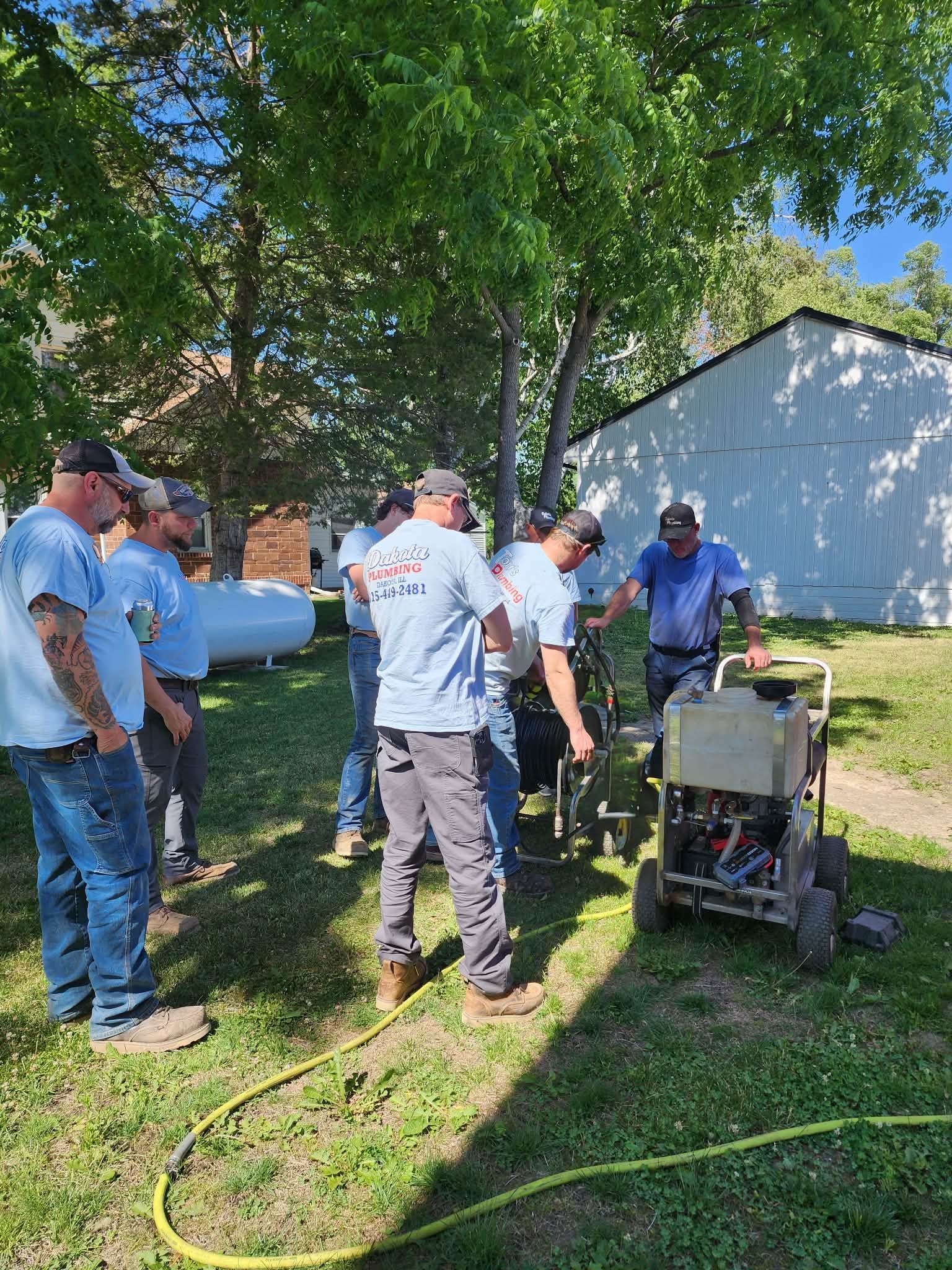 Several workers in white shirts gather around equipment in a shaded yard near a house