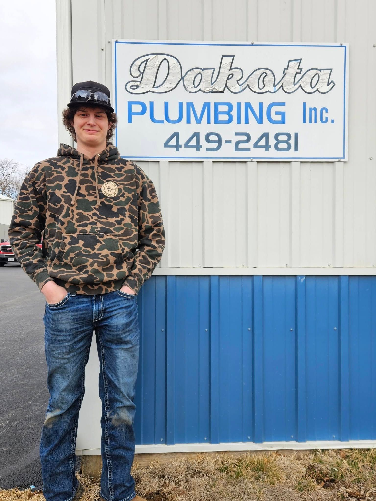 Person standing beside a sign for Dakota Plumbing Inc. with blue and white siding in the background.