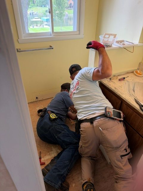 Two workers kneel on a kitchen floor, repairing wood beside a countertop and window.