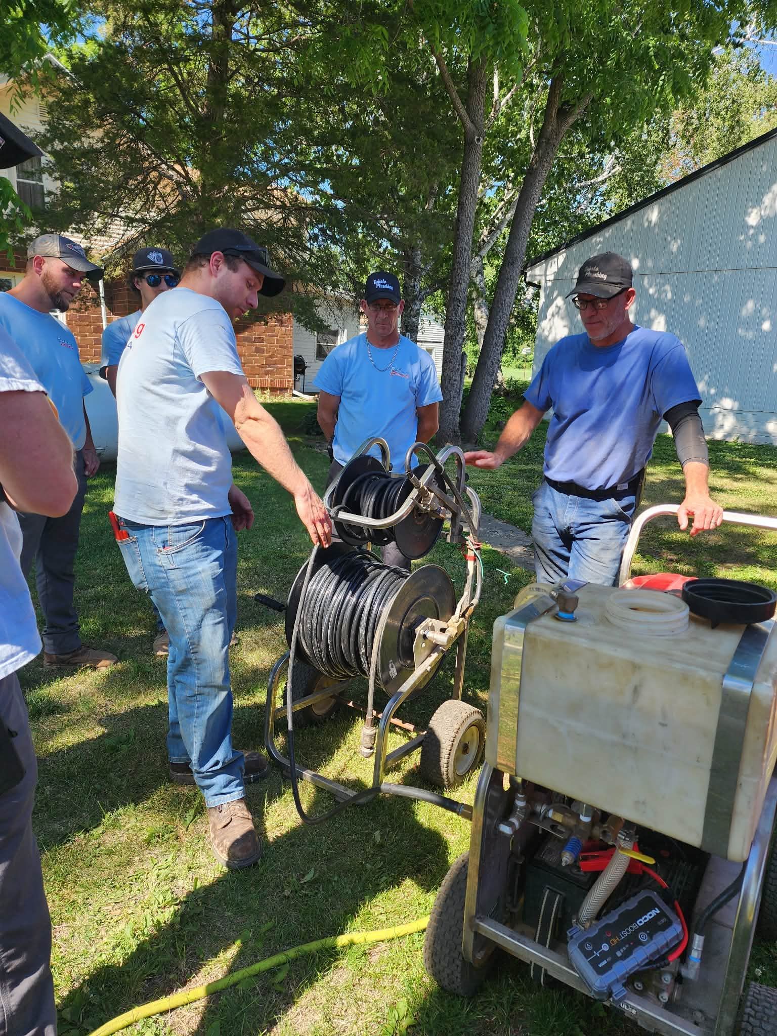 Men gathered outdoors around a small machine and cart under trees, discussing and handling equipment.