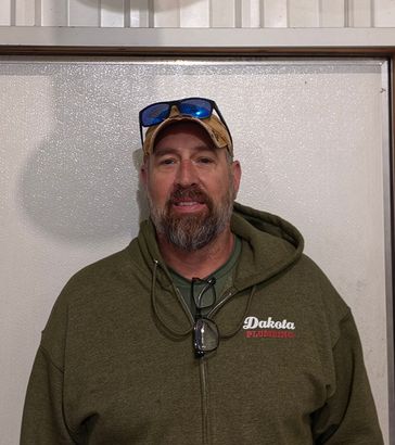 Man in green hoodie and tan cap standing indoors against a white wall.