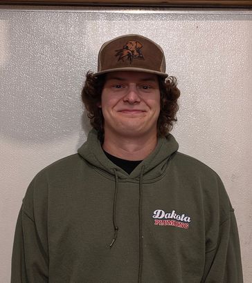 Person in a green hoodie and brown cap stands against a light textured wall, smiling.