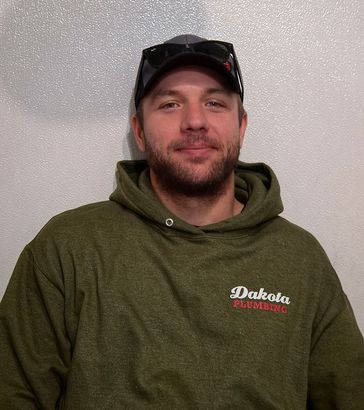 Person in an olive green hoodie and black cap, smiling against a plain light wall.