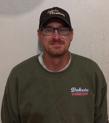 Man wearing a black cap, glasses, and an olive sweatshirt with a Dakota print, standing against a plain wall.