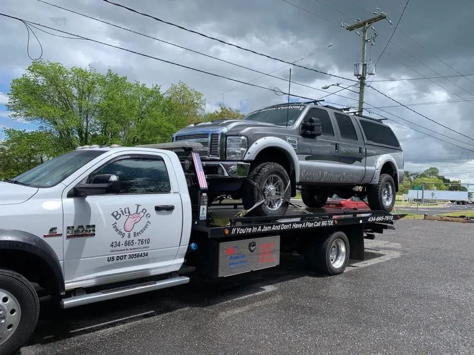 Tow truck transporting a large gray SUV on a street. Truck has business logo. Overcast sky in the background.