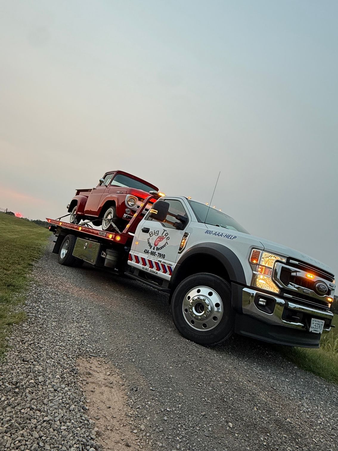 Tow truck hauling a red car on a gravel road at dusk.
