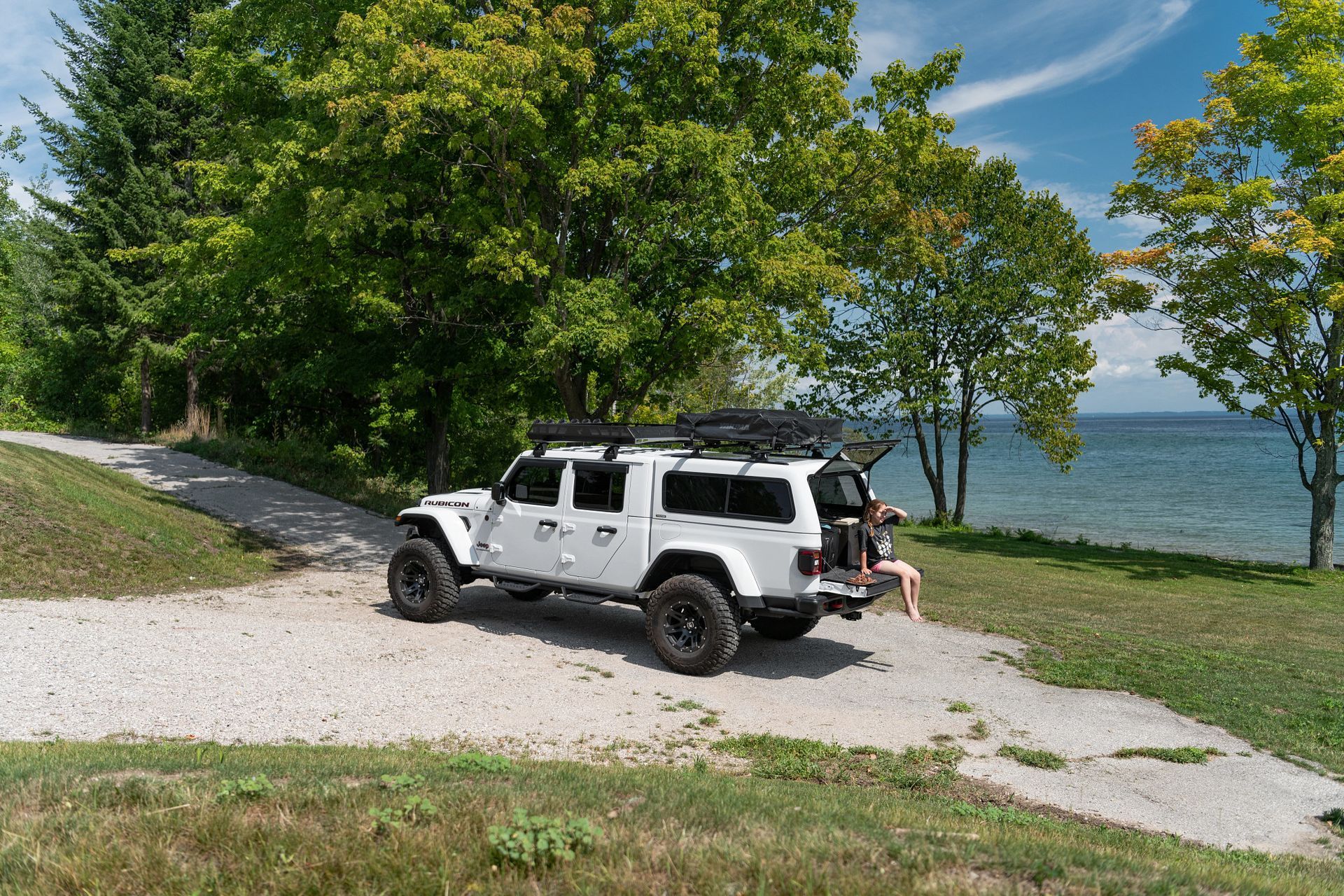 A white Jeep truck parked on a gravel path by a lake with its tailgate open and a person sitting nearby.