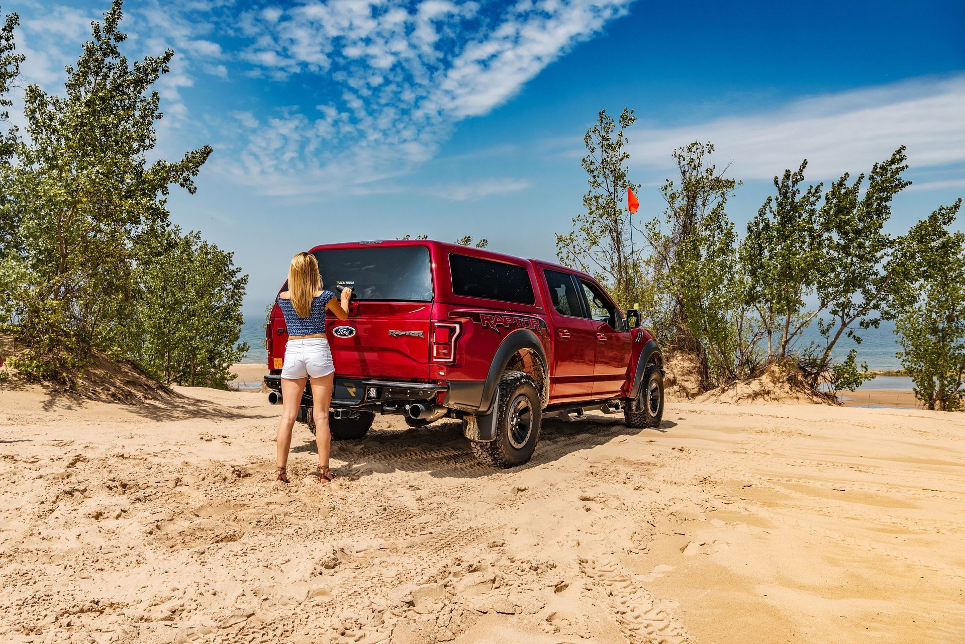 A person stands by the back of a red pickup truck with a topper, parked on a sunny, sandy dune surrounded by trees.