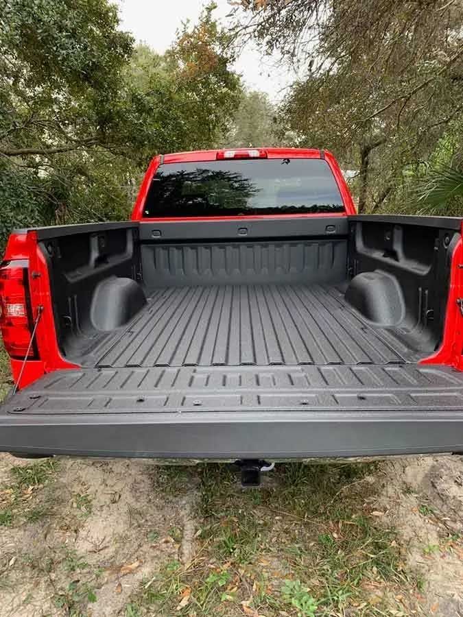Red pickup truck bed, open, lined with black, against a backdrop of trees.