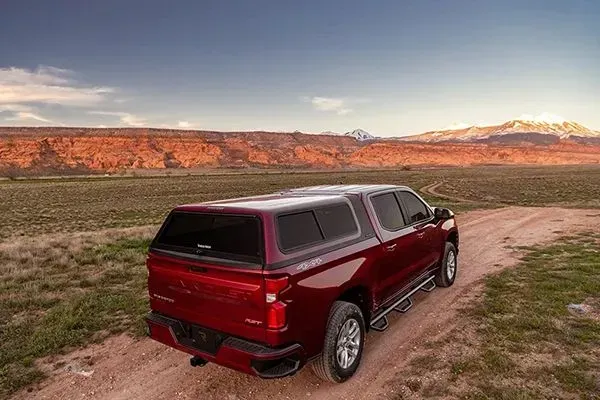 A dark red pickup truck with a camper shell parked on a dirt path in a scenic desert landscape at sunset.