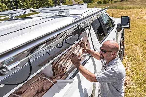A person in sunglasses retrieves long, wooden boards from the open, side-loading storage compartment of a white truck.
