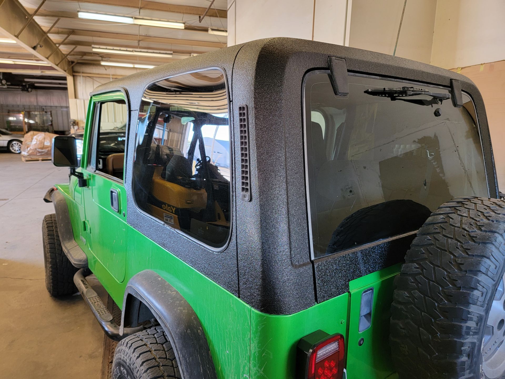 Green Jeep Wrangler with black hardtop and spare tire in a garage.