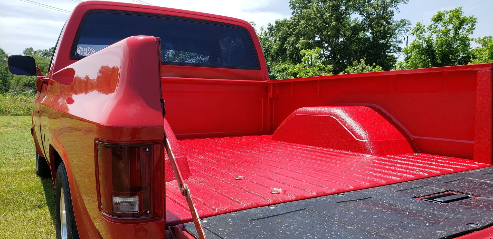 A red pickup truck with a vibrant, freshly painted bed and a black rubber liner, parked in a grassy field.