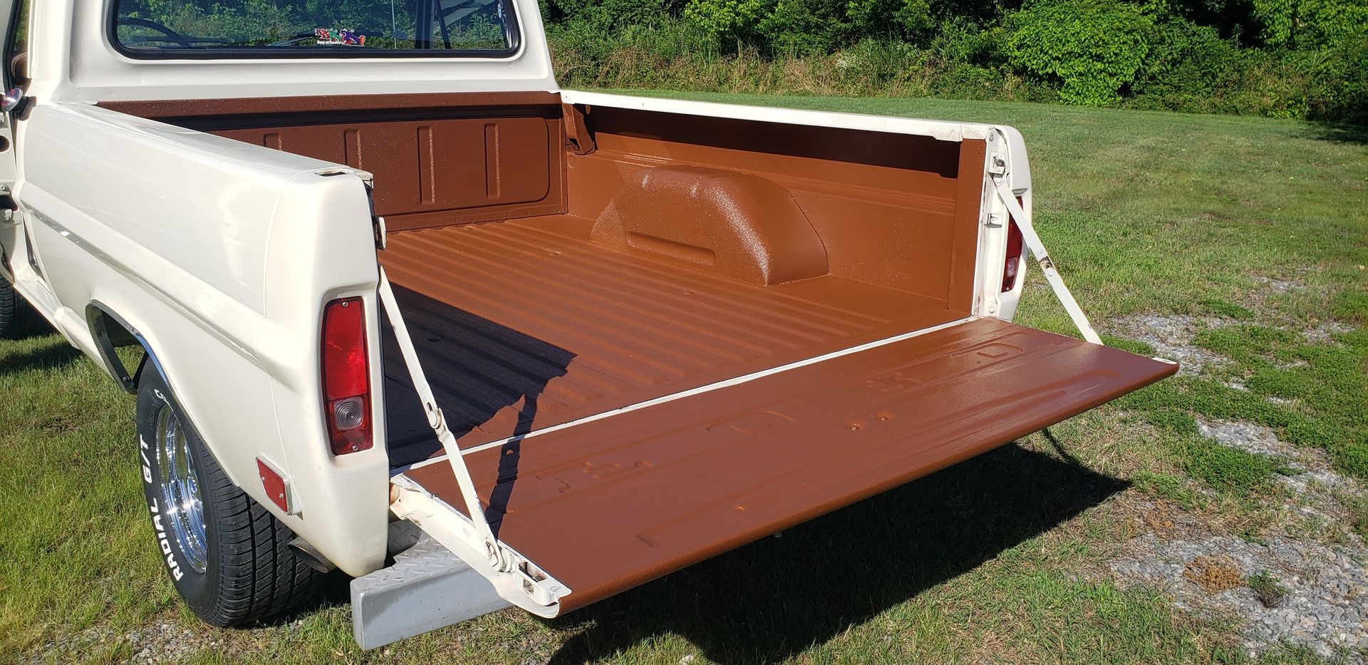A cream-colored pickup truck with its tailgate down, revealing a painted brown truck bed in a grassy field.