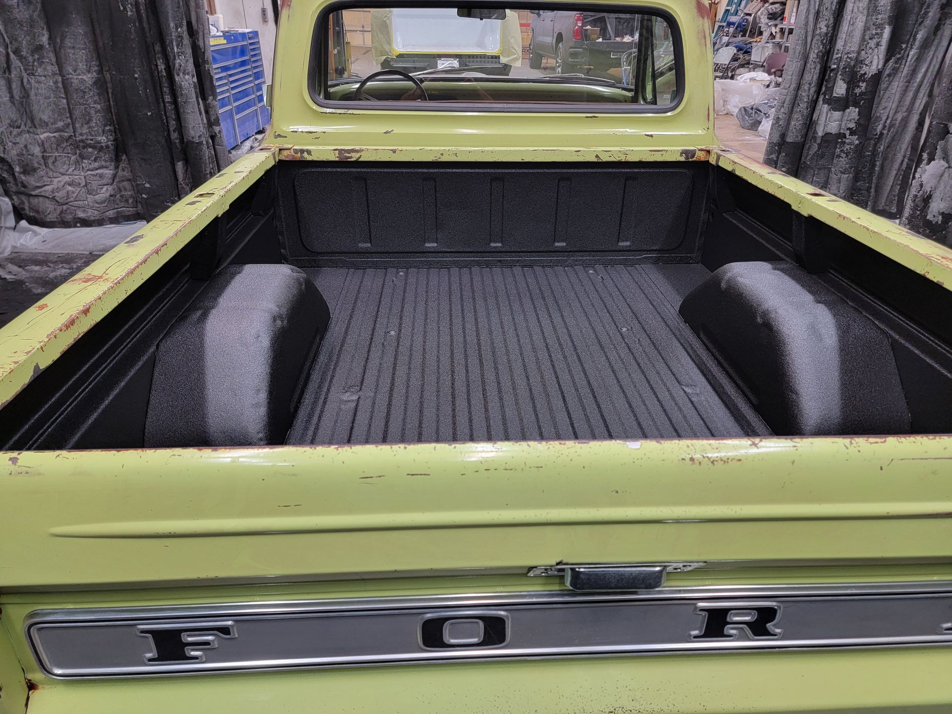 A pale green vintage Ford pickup truck bed with a black textured liner, viewed from the rear.
