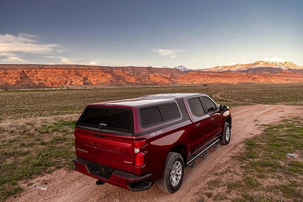 A red pickup truck with a camper shell parked on a dirt path in a desert landscape with red cliffs and distant mountains.