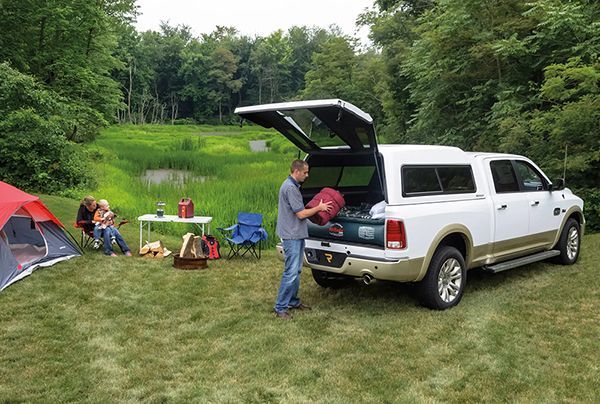 A person packing a white truck near a campsite with a red tent, a folding table, and a green meadow in the background.