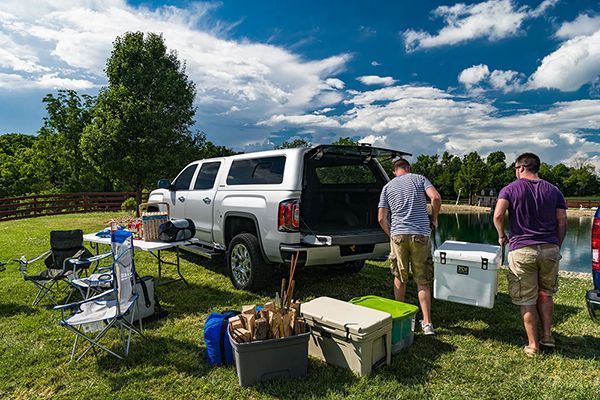 Two people unload camping gear and a cooler from a silver pickup truck parked by a lake on a sunny, cloudy day.