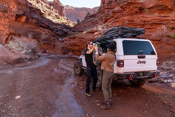 Two people stand by a white Jeep in a desert canyon, adjusting gear near the rear of the vehicle.