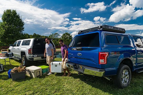 Two people unload gear and coolers from a white truck and a blue pickup truck on a grassy field under a cloudy blue sky.