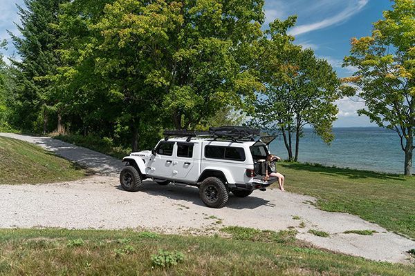 A white Jeep Gladiator with a camper shell parked on a lakeside gravel path, with a person sitting on the open tailgate.