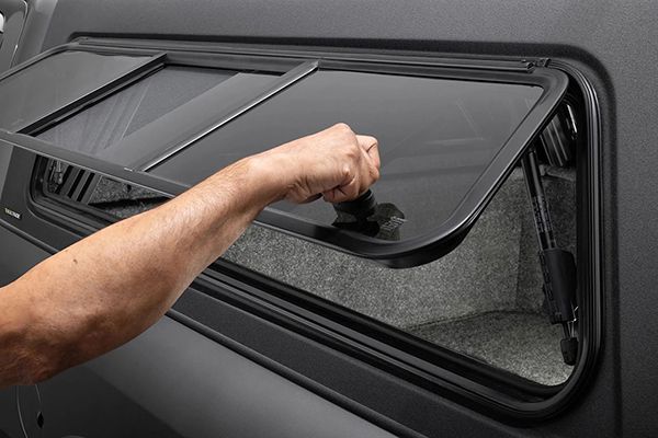 A hand opening a side-access window on a black truck cap with a carpeted interior.