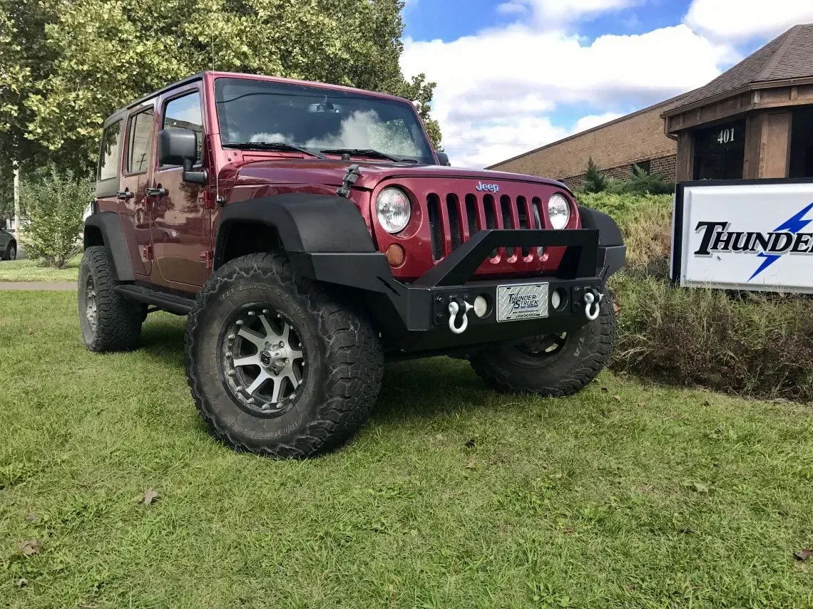 Red Jeep Wrangler parked on grass, black fender flares, custom bumper, large tires, and logo sign.