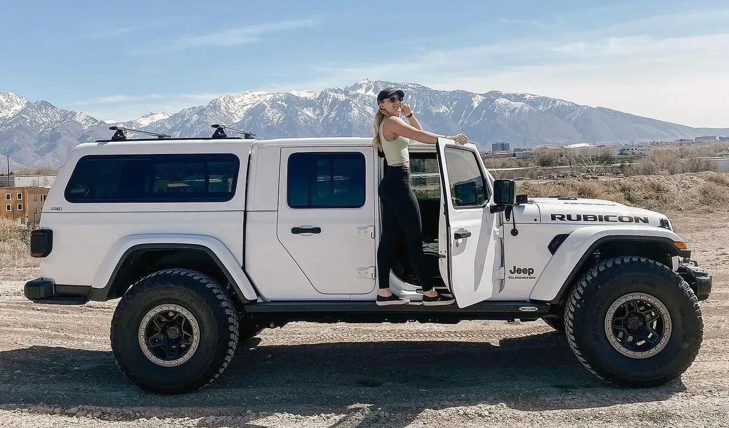 Woman standing in white Jeep, open door. Mountains in background. Sunny day.