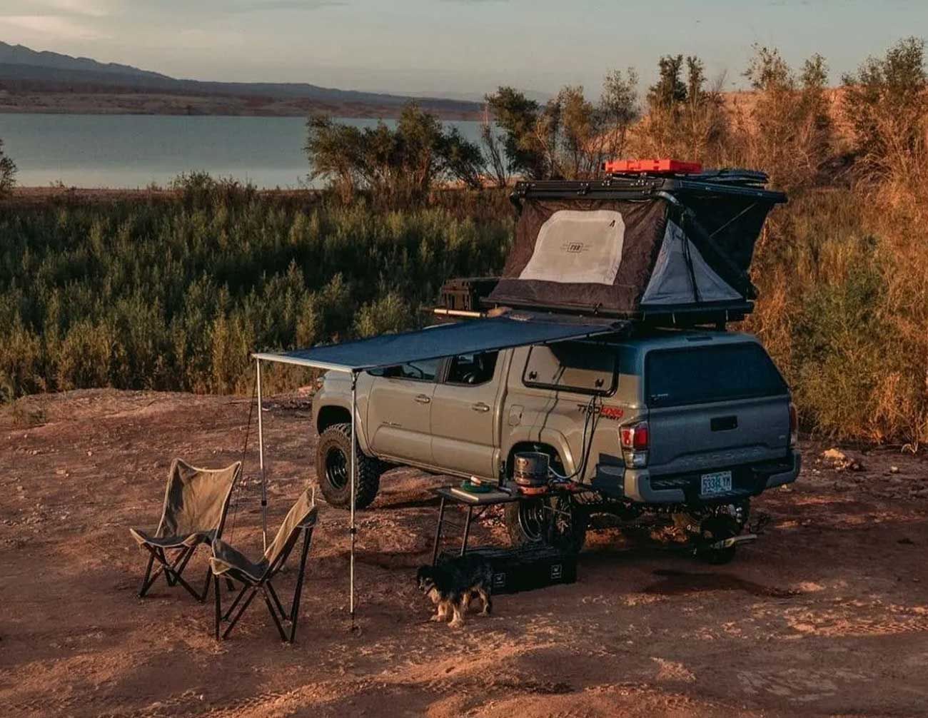 Overland truck with rooftop tent, awning, camping chairs, and a dog, by a lake.