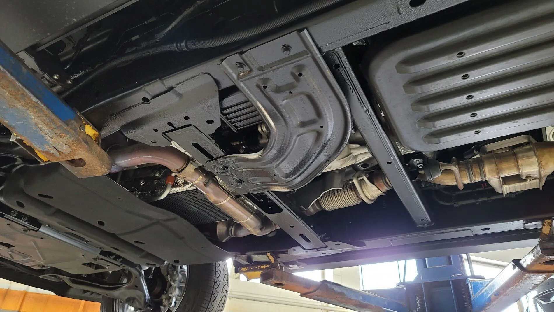 Undercarriage of a vehicle raised on a lift; shows exhaust pipes, metal frame, and protective plates.