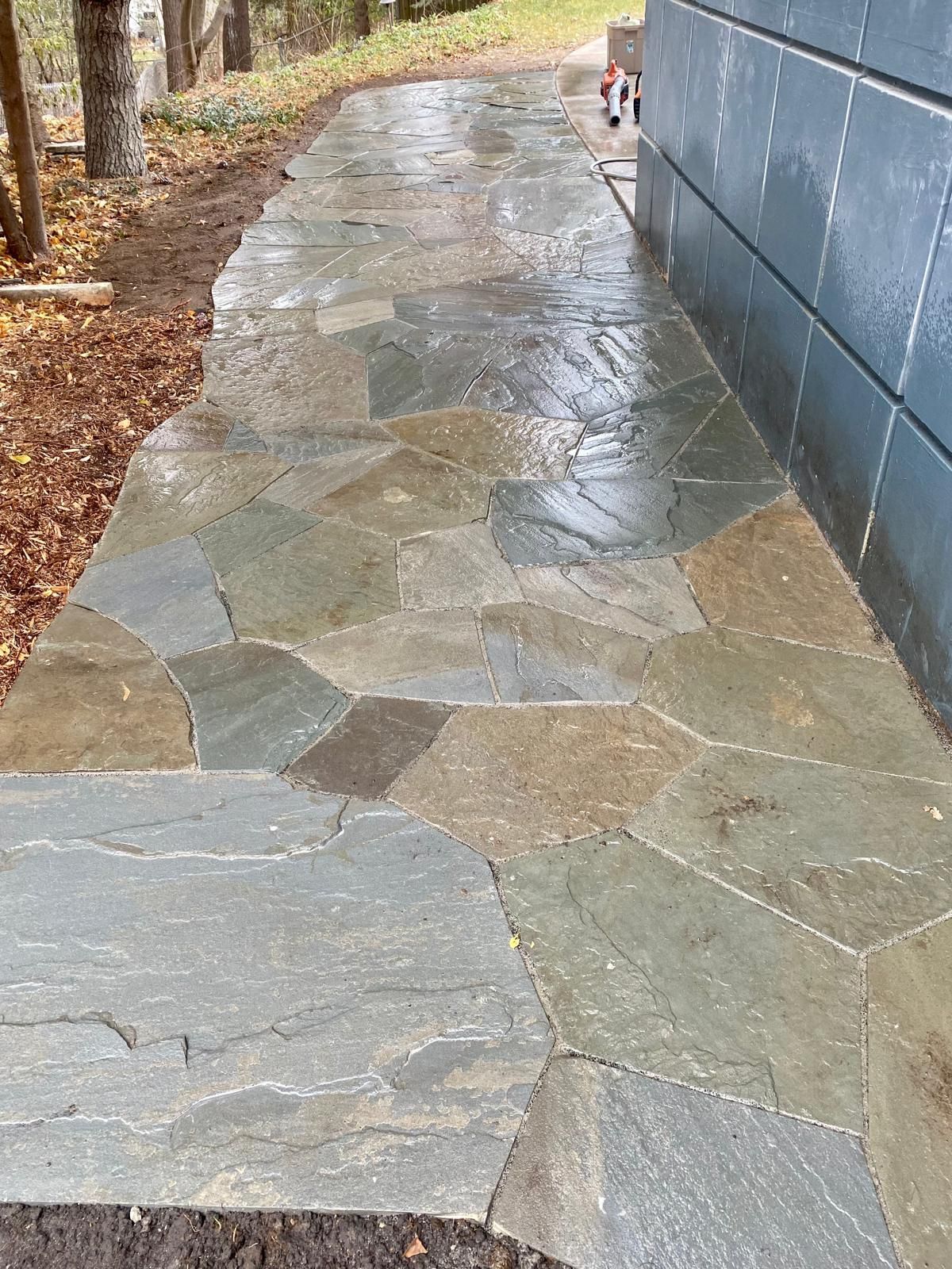 Flagstone walkway next to a dark blue building, surrounded by mulch and trees.