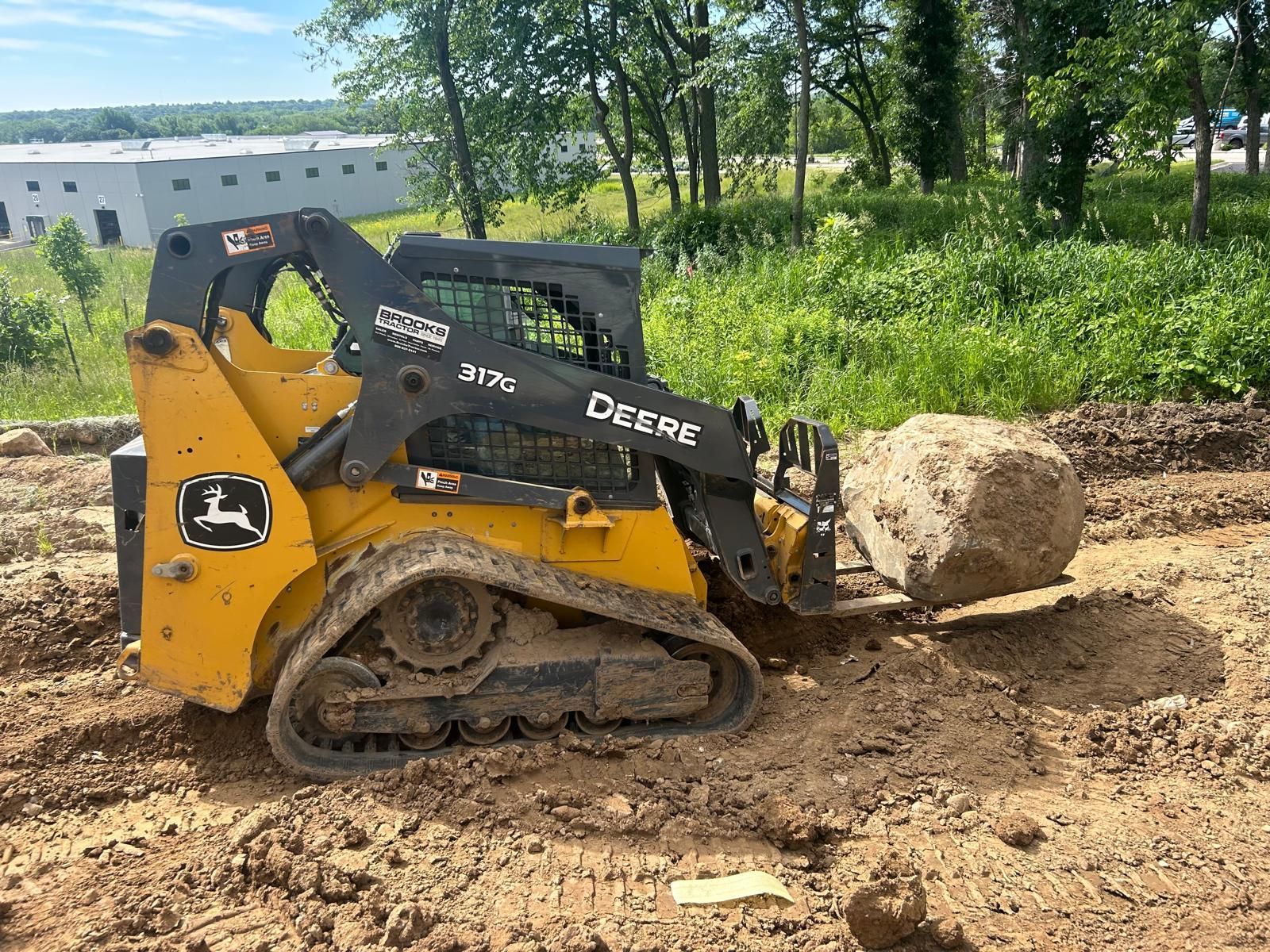 Yellow John Deere skid steer, lifting a large rock in a dirt setting, outdoors.