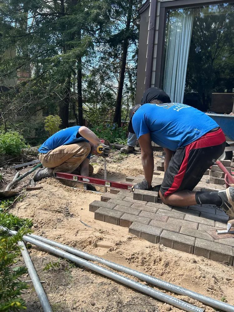 Two workers are installing pavers on a sandy base outdoors near a building. One uses a level.
