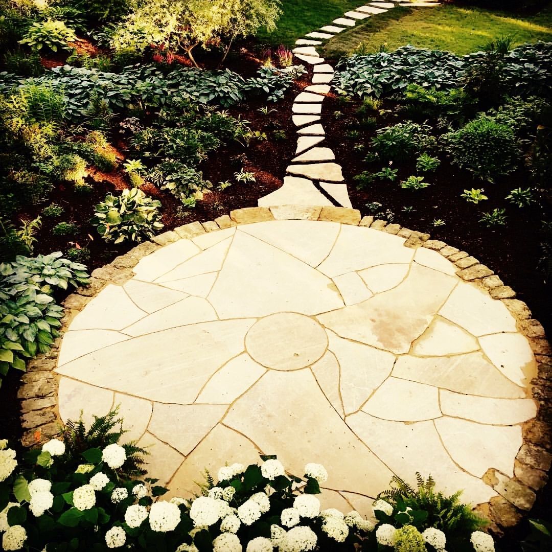A stone path leads to a circular patio surrounded by lush green plants and white flowers.