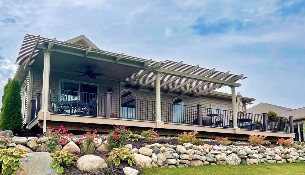 House with deck, pergola, and stacked stone retaining wall. Red flowers and green grass. Cloudy sky.