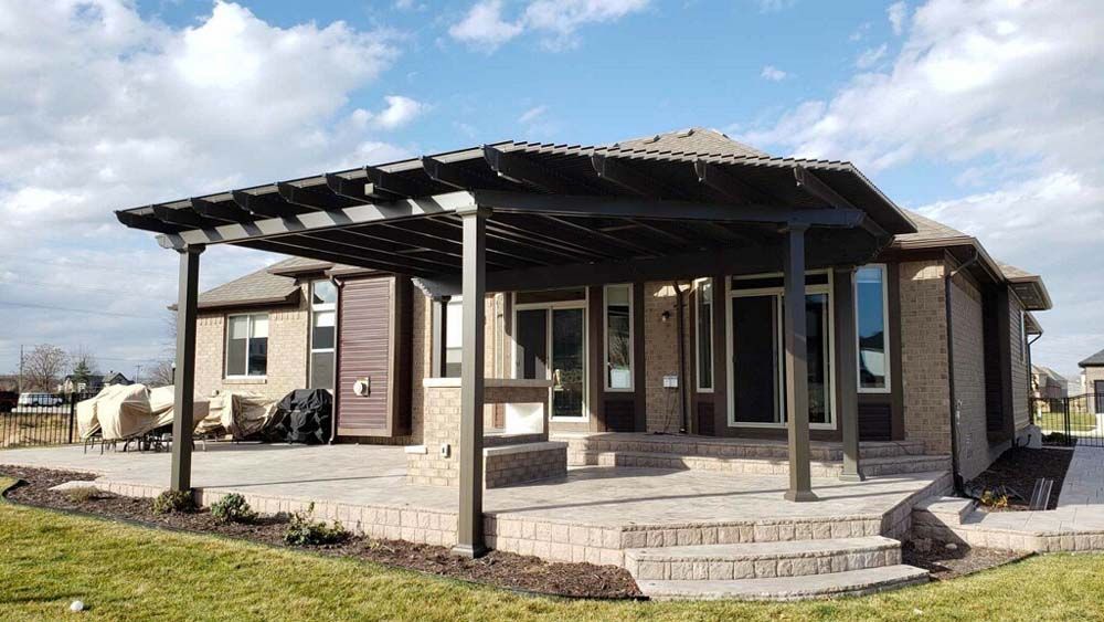 A dark brown pergola shades a brick patio outside a tan brick house on a sunny day.