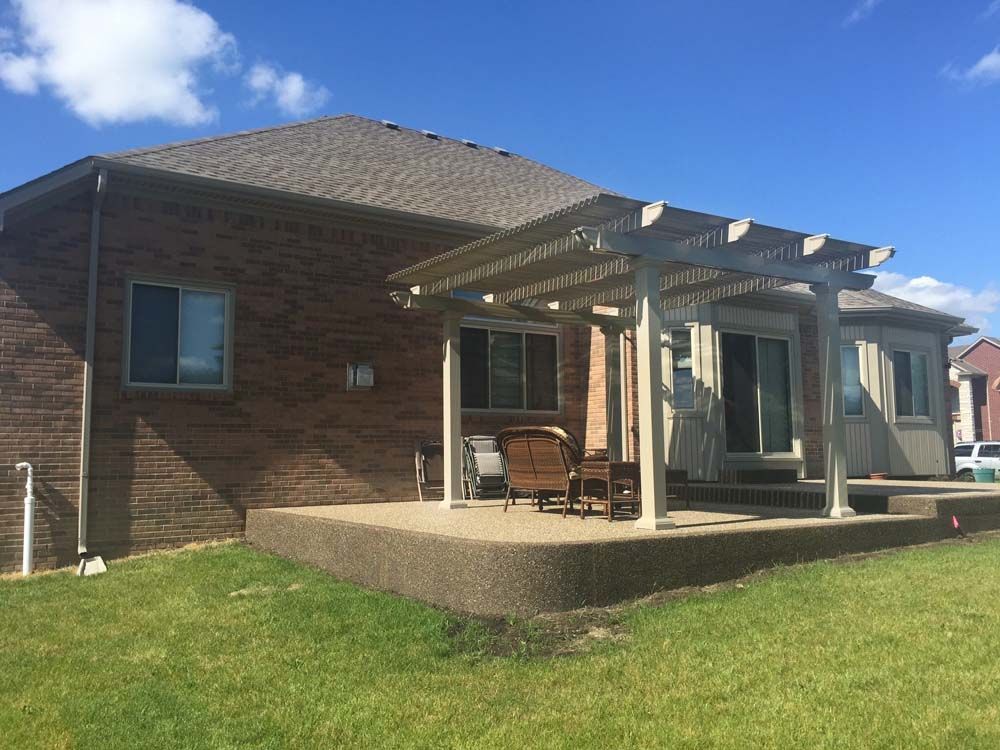 Brick house with concrete patio, pergola, and sliding glass doors. Green lawn and blue sky.