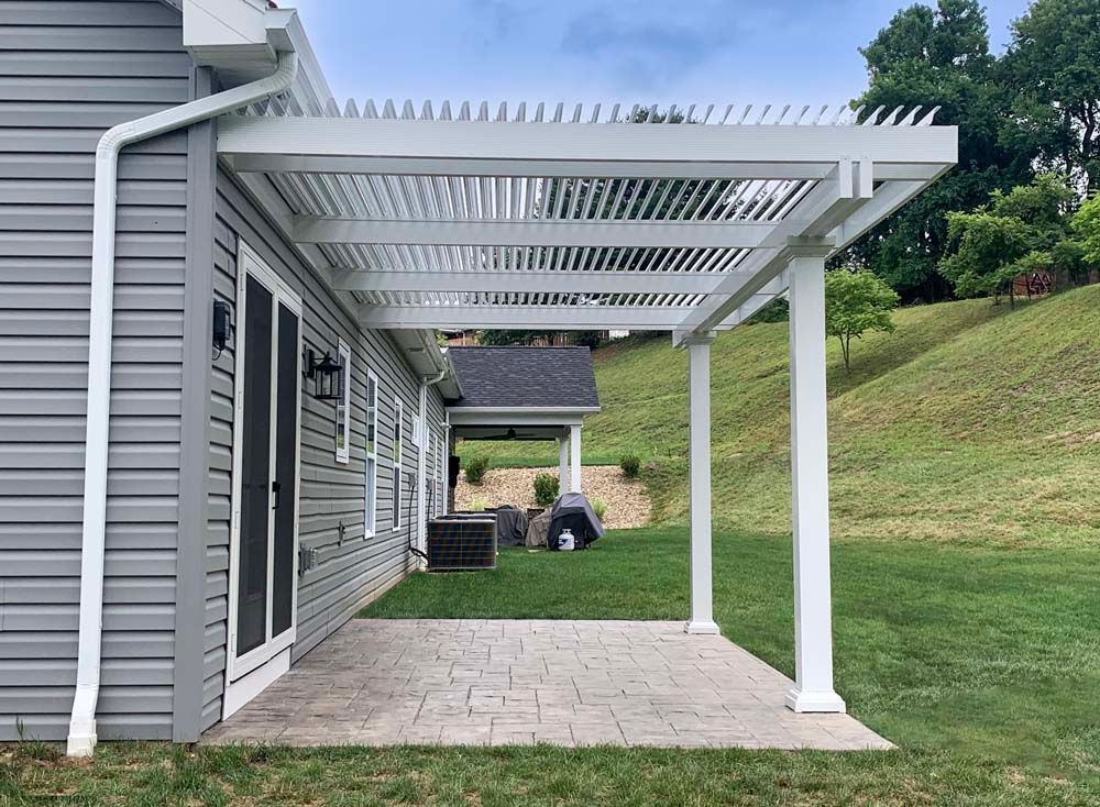 White pergola attached to a gray house, over a brick patio and green lawn.