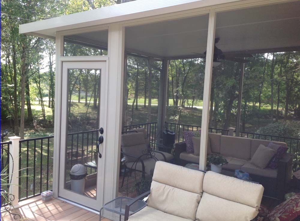 Screened porch with seating, door, and view of trees. Cream-colored frame, brown decking.