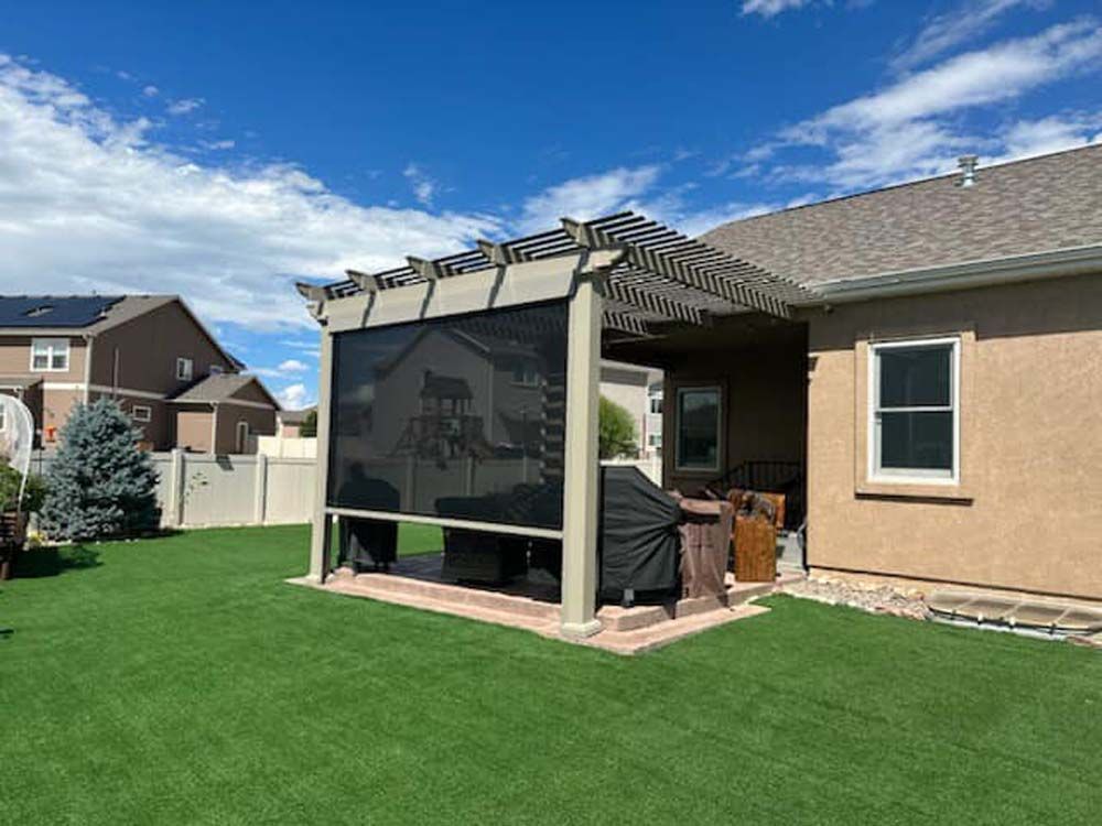 Pergola with black shade on a patio with grill, on green grass lawn, against a house and blue sky.