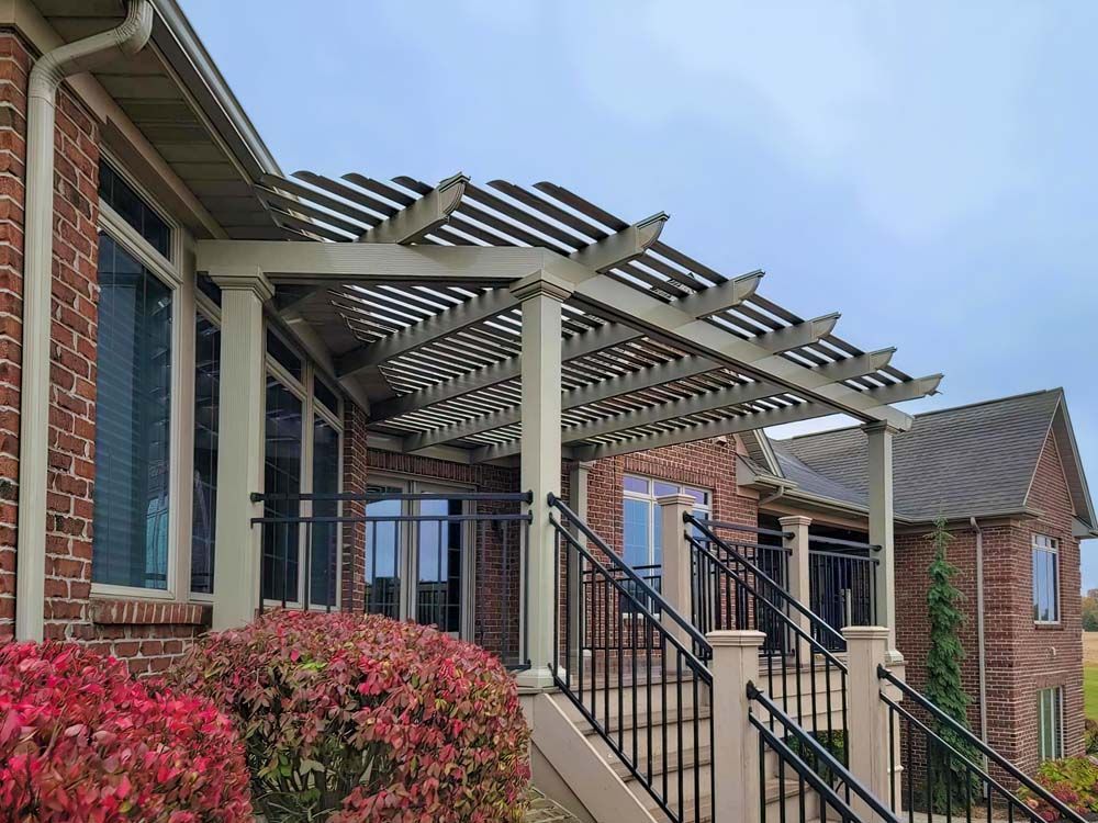 Pergola over a brick house patio with stairs, black railing, and red bushes. Cloudy sky.