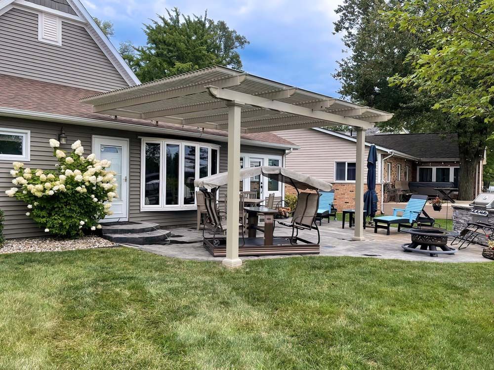 Backyard patio with pergola, table, chairs, and house. Green grass and blue sky.