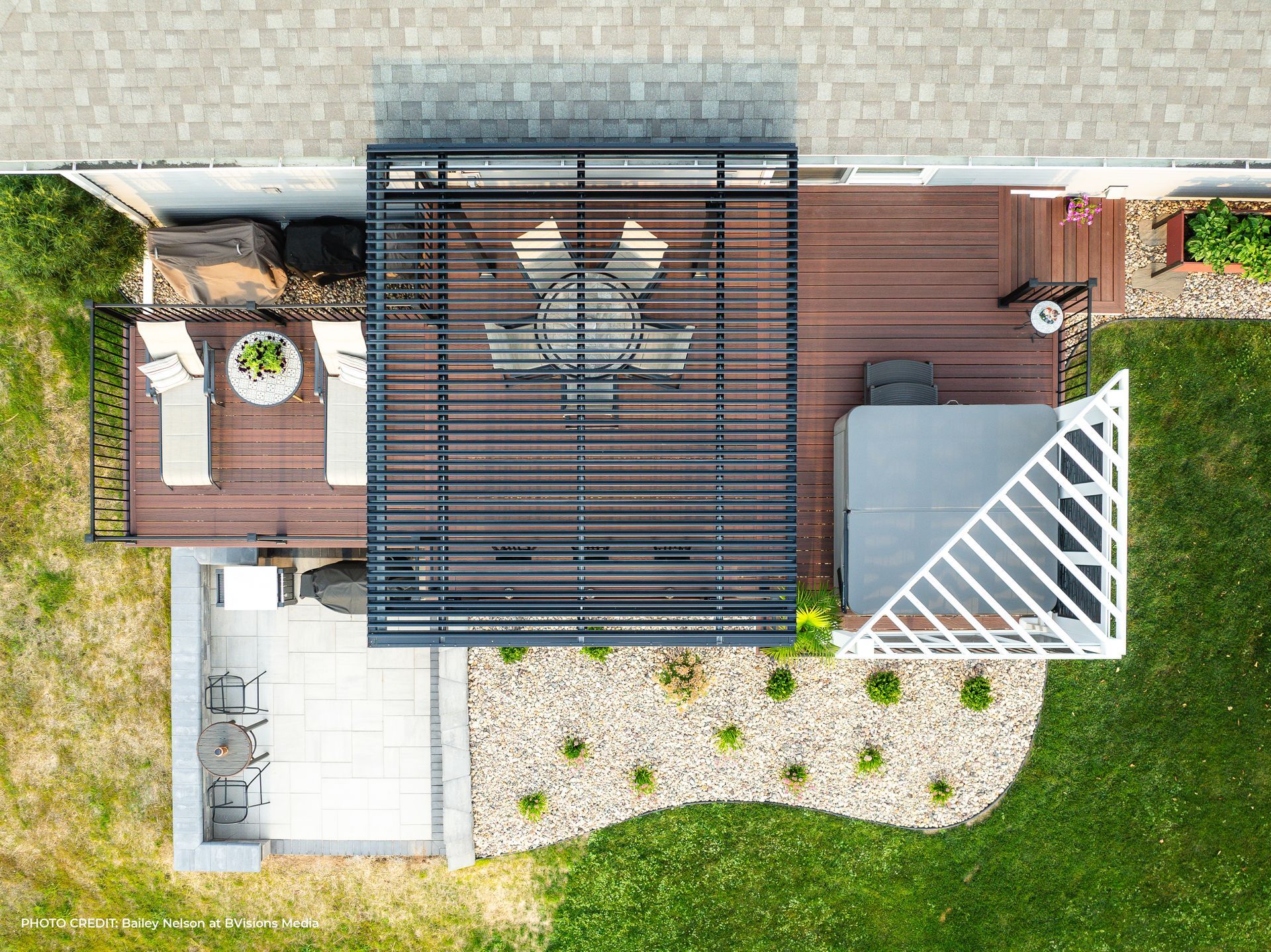 Aerial view of a backyard deck with a dark pergola, patio furniture, a hot tub, and a small stone patio area.
