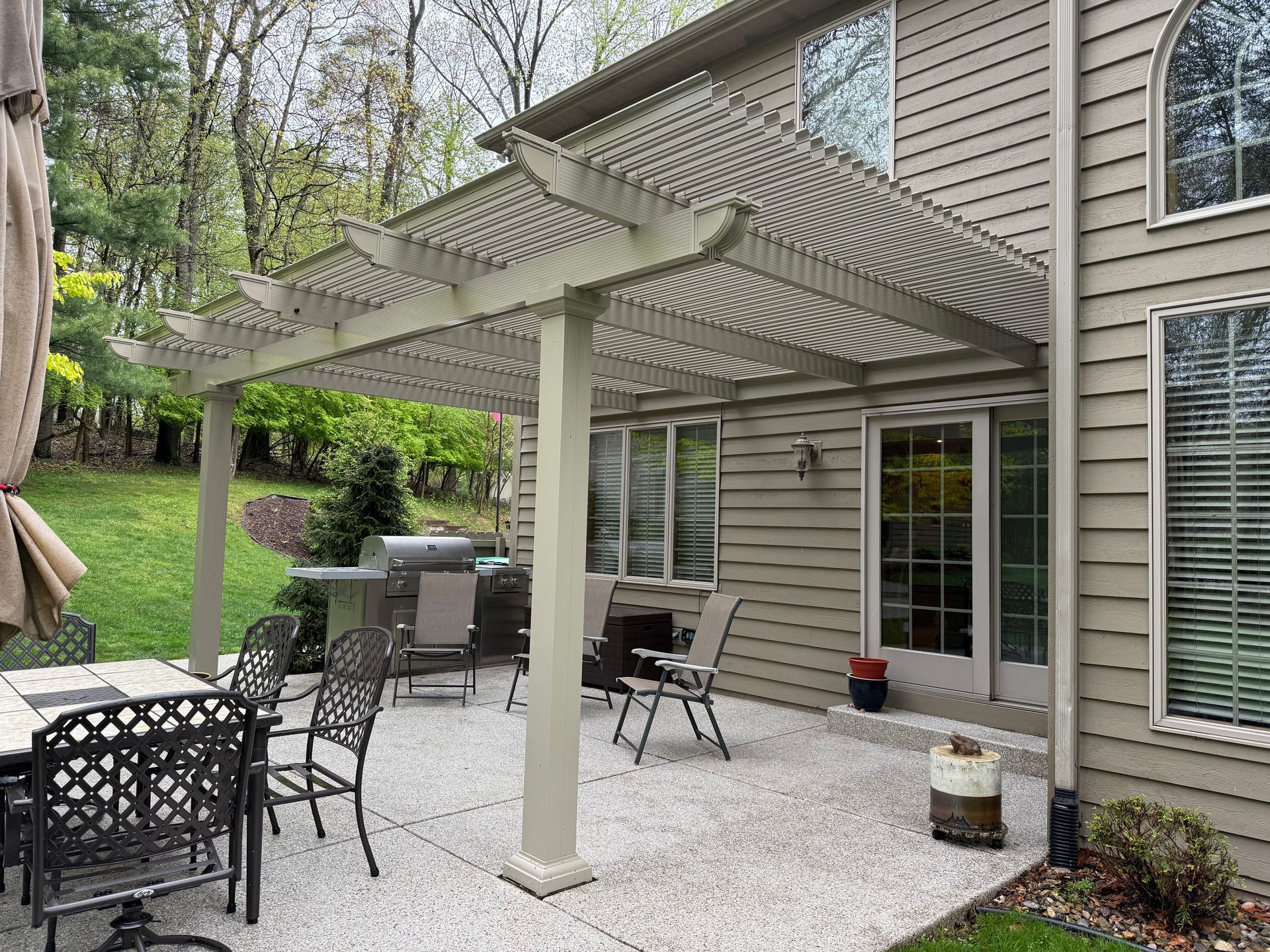 A beige wooden pergola shades a stone patio with outdoor dining furniture and a grill next to a house with beige siding.