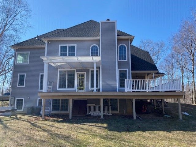 A two-story grey-sided house featuring a deck with a pergola, white railing, and a tall brick chimney against a blue sky.