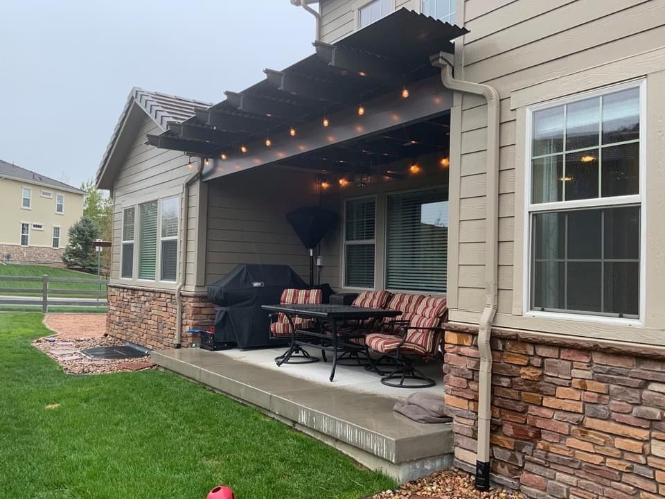 A covered outdoor patio with a dining table, chairs, grill, and string lights attached to the side of a tan house.