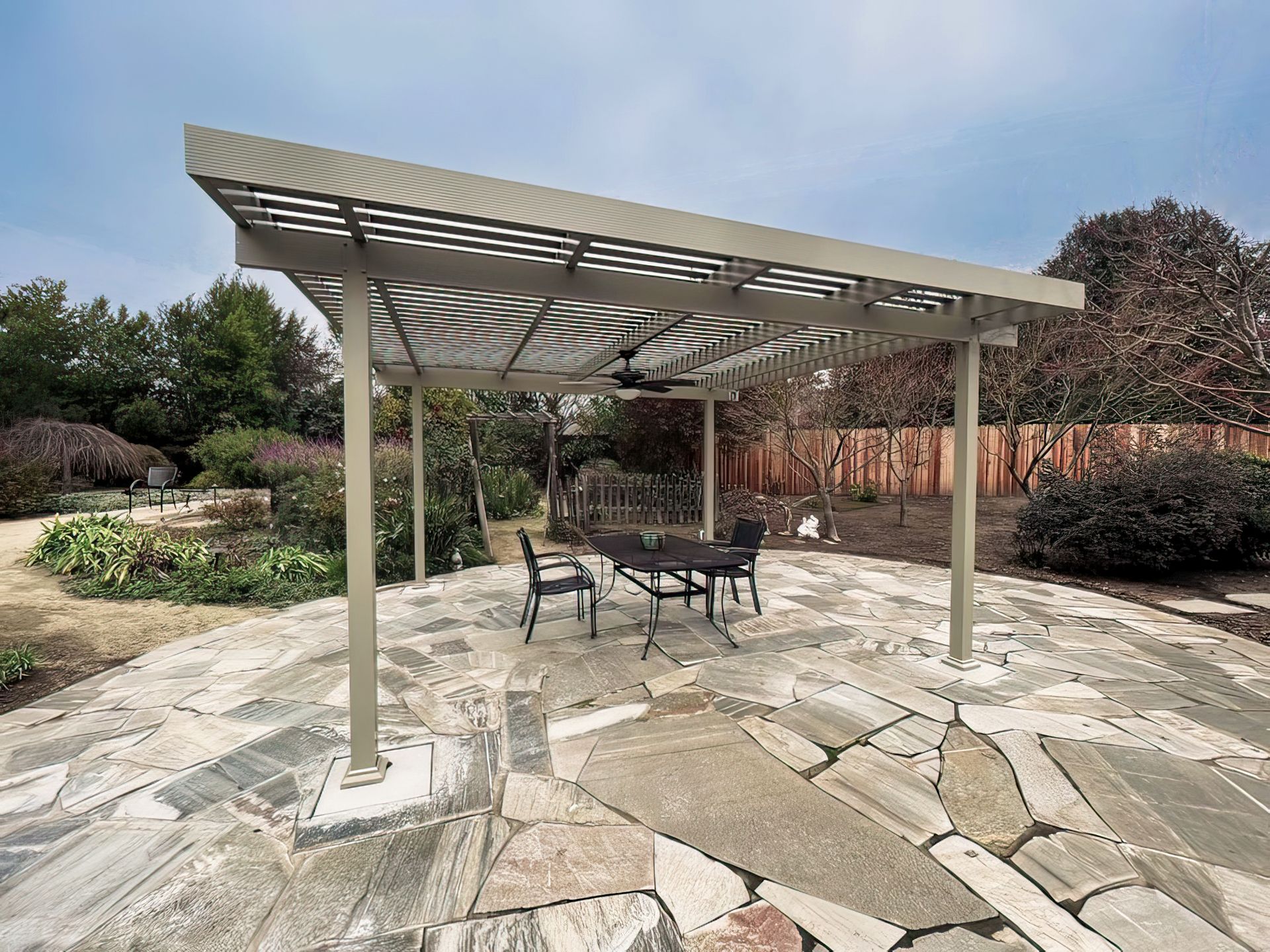 A white pergola attached to a grey house, featuring a slatted roof over a paved patio, overlooking a green grassy hill.