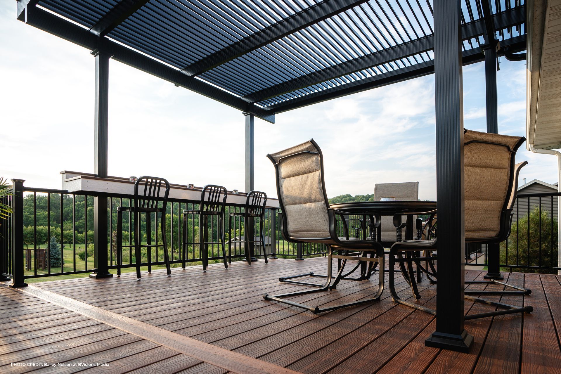An aerial view of a house deck featuring a black pergola, outdoor furniture, and a stone patio below in a grassy yard.