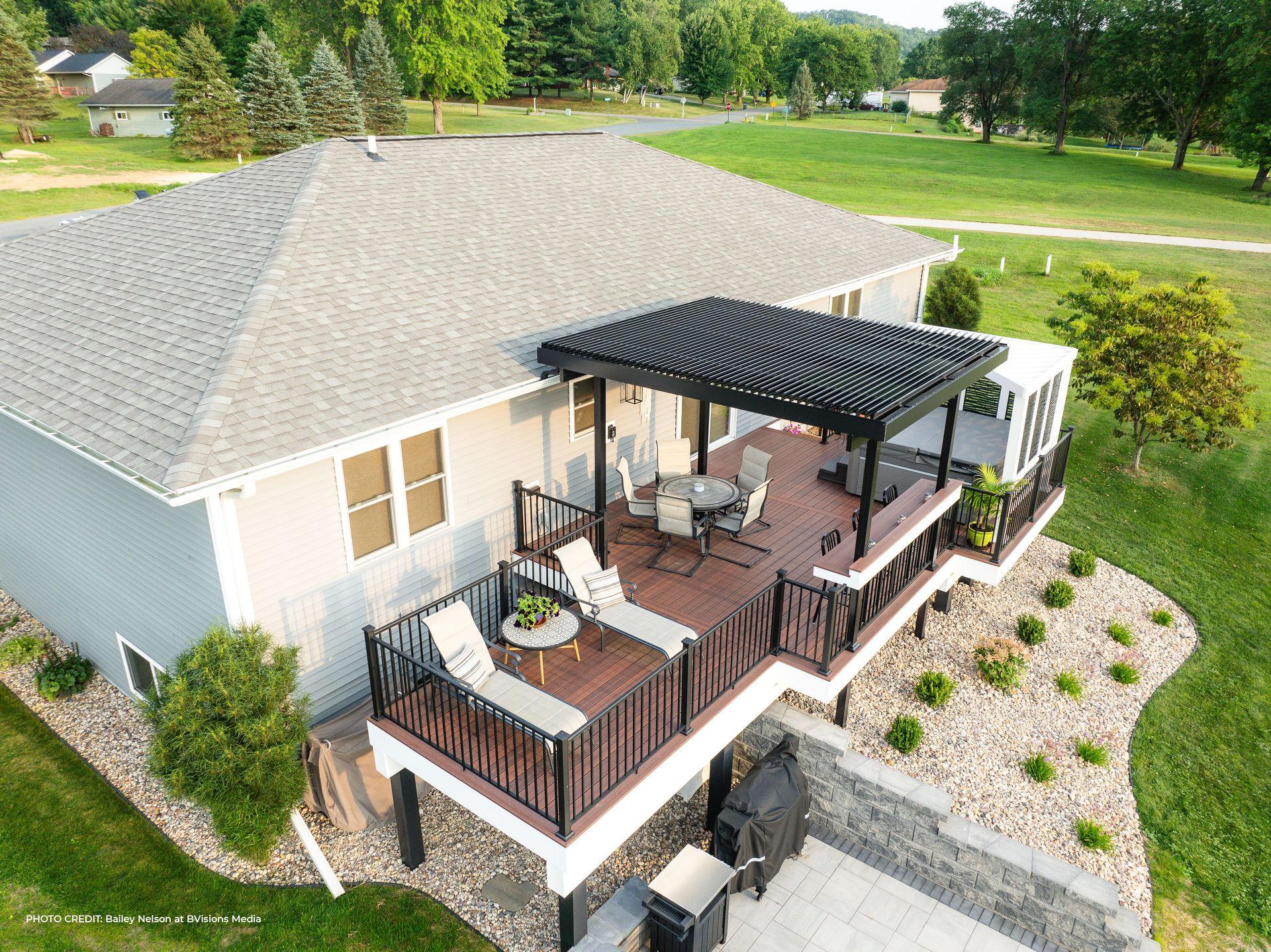 An elevated deck with a pergola, patio furniture, and a stone-lined backyard viewed from an aerial perspective.